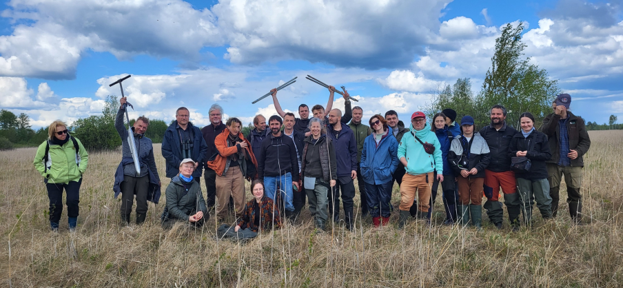 Grupowe zdjęcie członkiń i członków Centrum Ochrony Mokradeł z wycieczki terenowej podczas wyjazdowego walnego zgromadzenia w Urszulinie w 2025 roku. Osoby pozują na bagnie - większość stoi, część kuca, niektóre z osób trzymają części świdra torfowego, którym badana była miąższość torfu.